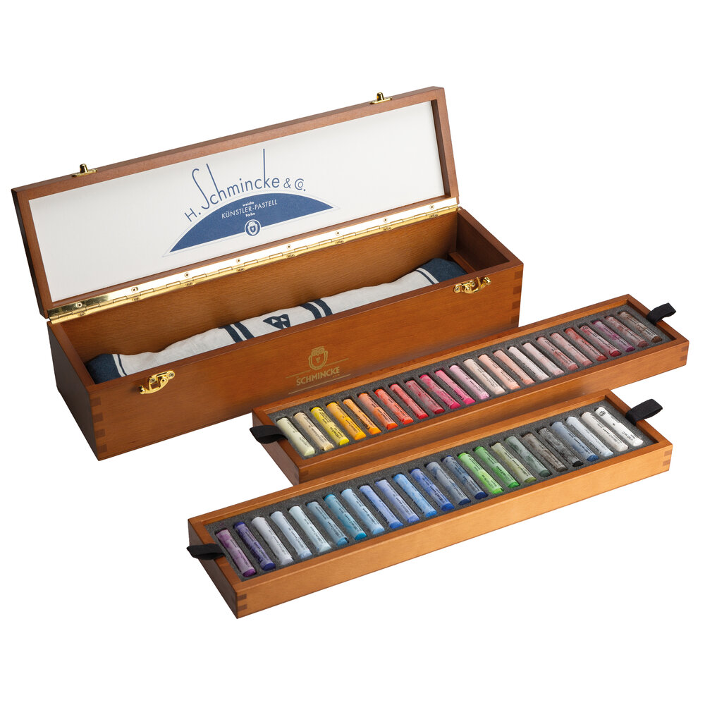 Geopende houten Schmincke pastelkist in hoekaanzicht. Beide uitneembare inlays met pastels liggen los naast de kist. In de bode