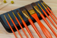 Sfeerfoto op een lichte, houten ondergrond. De 10 oranje Kangaro penselen met goudkleurige bussen en donker ponyhaar liggen uit